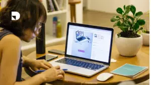 Una persona sentada viendo su computador y haciendo una lista en su cuaderno